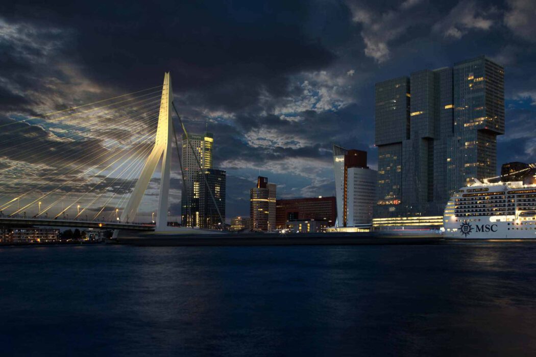 Die nachts beleuchtete weiße Erasmusbrücke vor dramatisch bewölktem Himmel; daneben moderne Hochhäuser mit vielen Lichtfenstern und ein großes Kreuzfahrtschiff am Ufer.