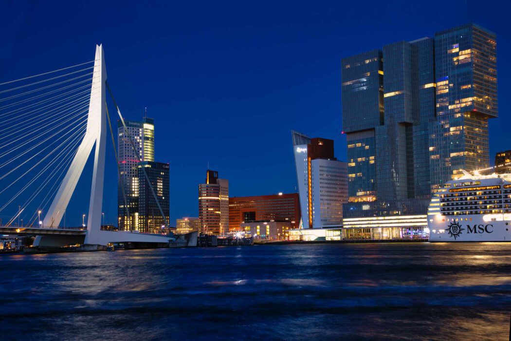 Die moderne, nachts beleuchtete Erasmusbrücke in Rotterdam, mit markantem weißem Pylon; im Vordergrund gehen zwei Personen unter einer Straßenlaterne, dahinter leuchten die Gebäude der Stadt.