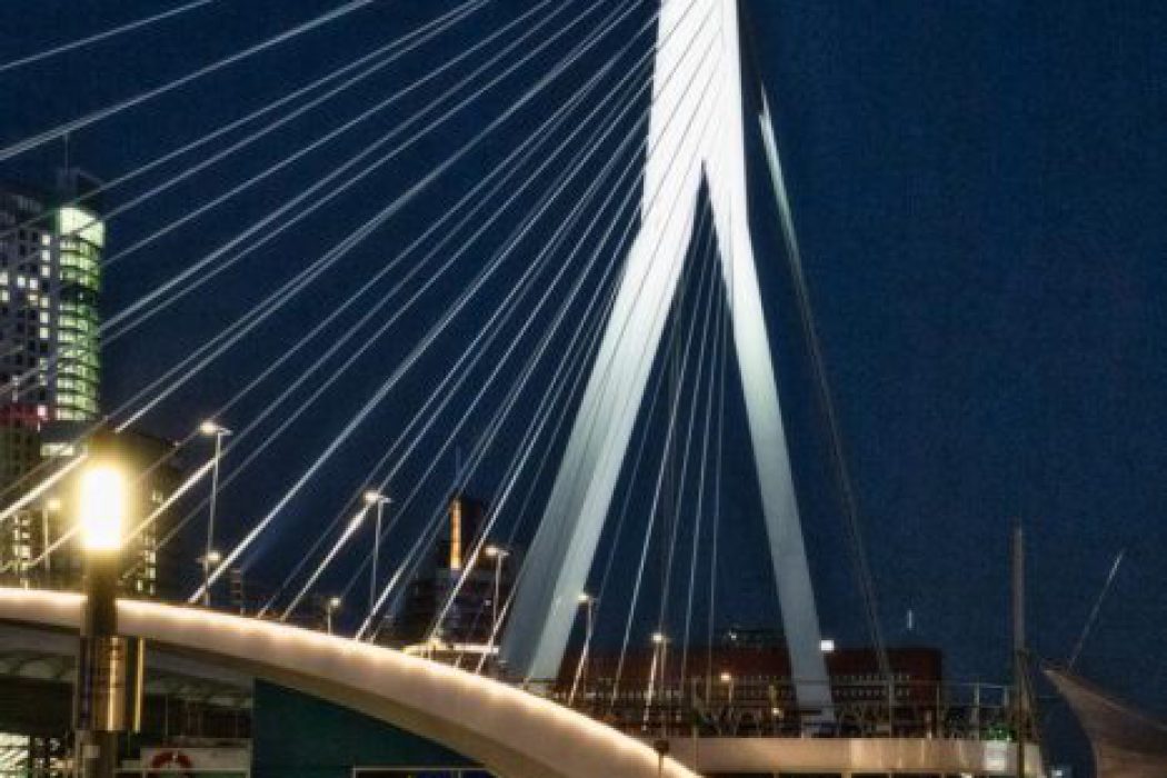 Eine moderne, nachts beleuchtete Schrägseilbrücke mit markantem weißem Pylon; im Vordergrund gehen zwei Personen unter einer Straßenlaterne, dahinter leuchten die Gebäude der Stadt.