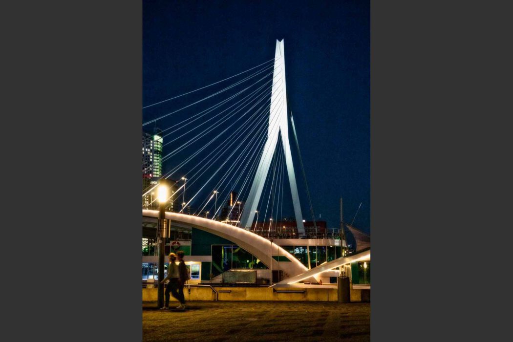 Nachtaufnahme der weiß beleuchteten Erasmusbrücke in Rotterdam. Der schräg geneigte Pylon ragt in den dunkelblauen Nachthimmel, die Stahlseile zeichnen feine Linien. Im Vordergrund gehen zwei Personen leicht unscharf an einer Straßenlaterne entlang der Maas-Promenade. Im Hintergrund beleuchtete Hochhäuser und ein Restaurantschiff am Ufer.