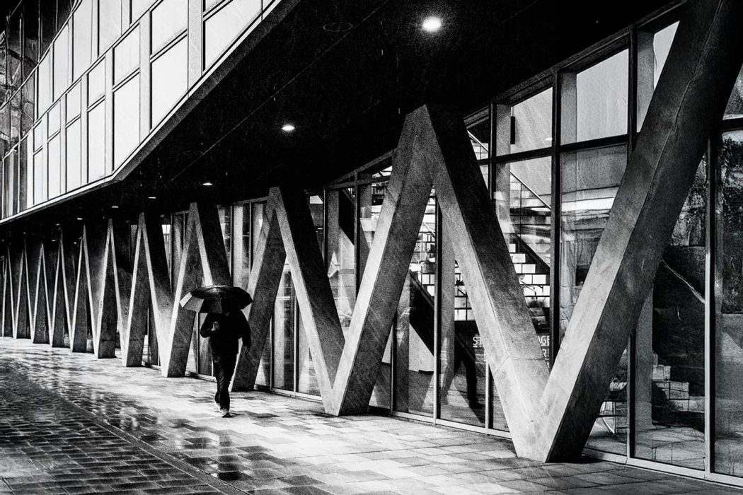Schwarz-Weiß-Aufnahme eines modernen Gebäudes mit diagonalen Betonpfeilern und großen Glasfenstern. Eine Person mit Regenschirm geht auf dem nassen Gehweg unter dem Vordach entlang.