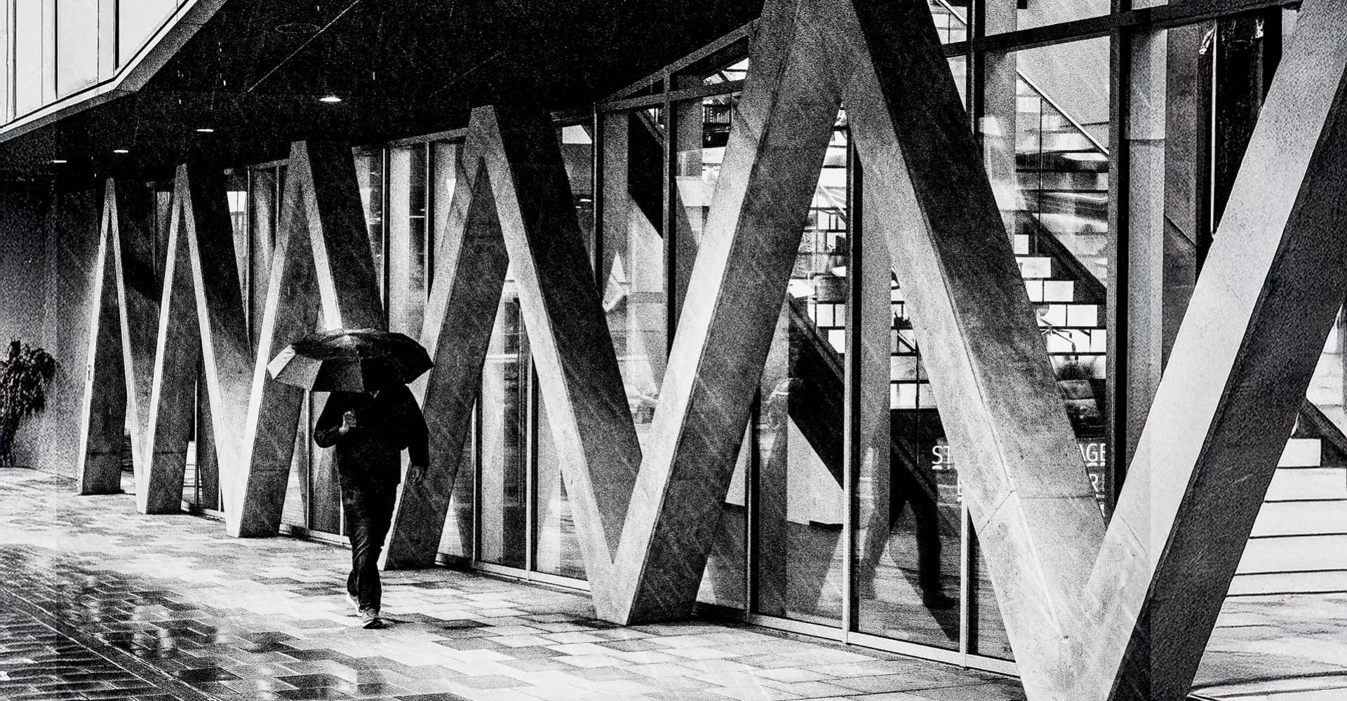 treetfotografie in Schwarz-Weiß: Person mit Regenschirm geht an einer modernen Fassade mit markanten A-förmigen Stützen entlang, spiegelnder nasser Boden.