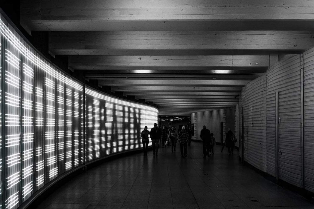 Schwarz-Weiß-Foto eines langen, geschwungenen U-Bahn-Gangs. Links erstreckt sich eine leuchtende Anzeigetafel, rechts eine geriffelte Wand. Mehrere unscharfe Personen gehen in der Mitte