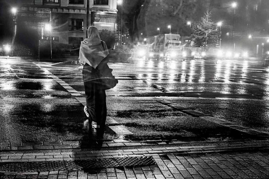 Person mit Kopftuch steht nachts an einer regennassen Kreuzung, Lichter spiegeln sich auf der Straße.