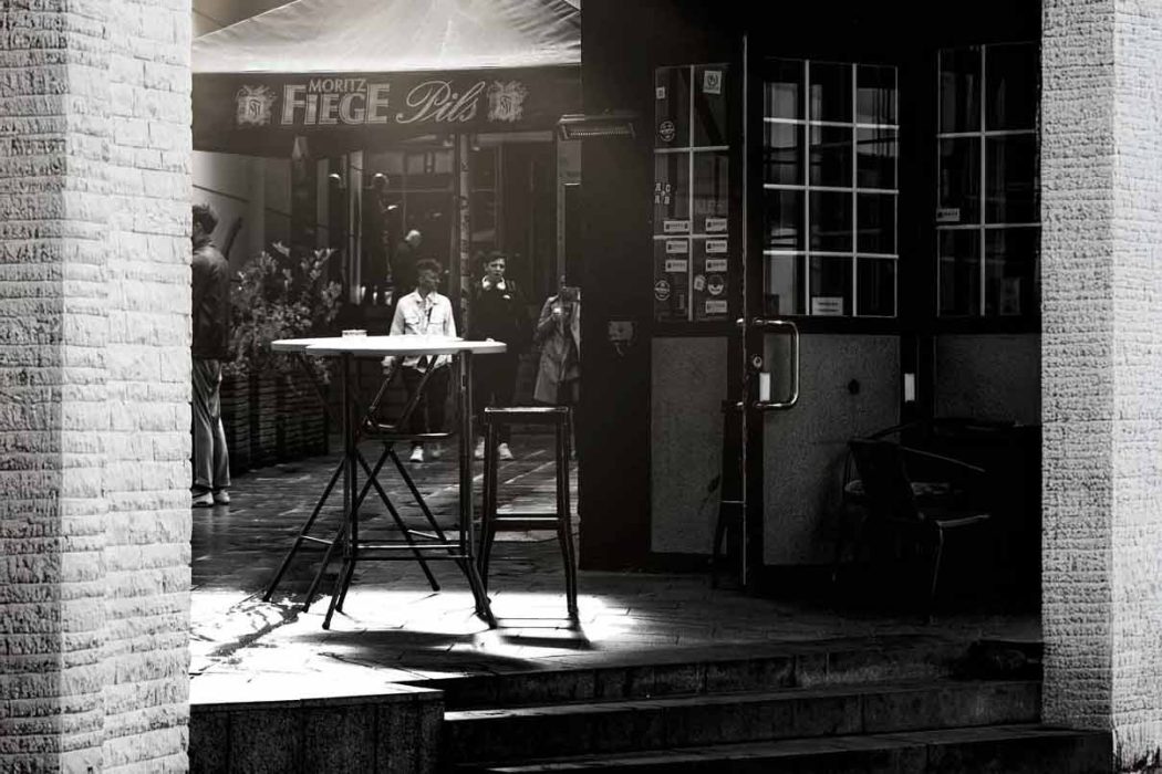 Außenbereich einer Bar mit Stehtisch und Menschen im Hintergrund, teilweise im Schatten