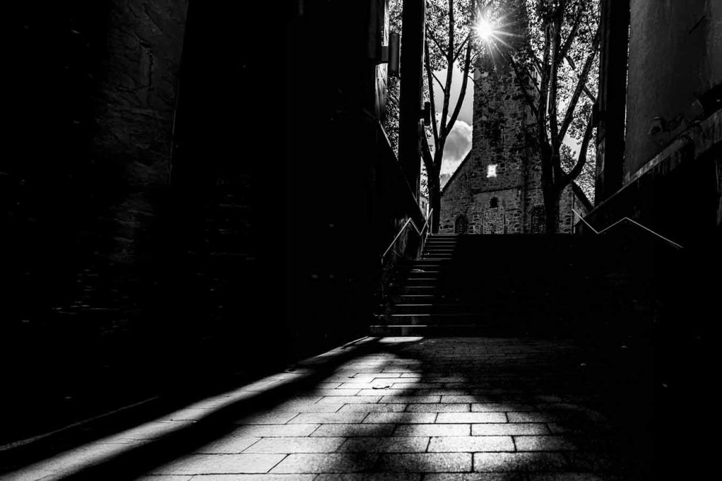 Schwarz-weiß Foto eines Treppenaufgangs mit Blick auf eine alte Kirche, Sonnenstrahlen scheinen durch Bäume.