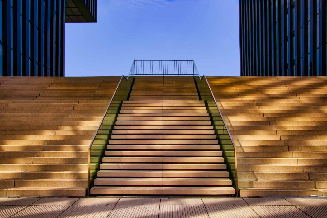 Hyatt Düsseldorf Treppe: Eine beeindruckende, breite Steintreppe führt zwischen zwei modernen Gebäuden des Hyatt Hotels in Düsseldorf nach oben, unter einem klaren blauen Himmel. Das Spiel von Licht und Schatten betont die architektonischen Linien. Ideal für Architekturfotografie, Reiseblogs über Düsseldorf und Hotelmarketing.