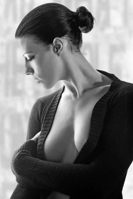 Ein Schwarz-Weiß-Porträt einer Frau in Seitenansicht. Sie hat ihre Haare zu einem Dutt hochgesteckt und trägt einen dunklen, offen sitzenden Strickpullover, der ihre Schultern und einen Teil ihres Dekolletés zeigt. Ihr Blick ist gesenkt, was eine kontemplative Stimmung erzeugt.