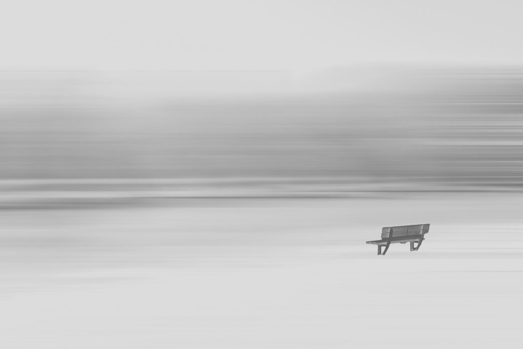 Holzbank in weiter, verschneiter Landschaft vor abstraktem, horizontal verschwommenem Hintergrund