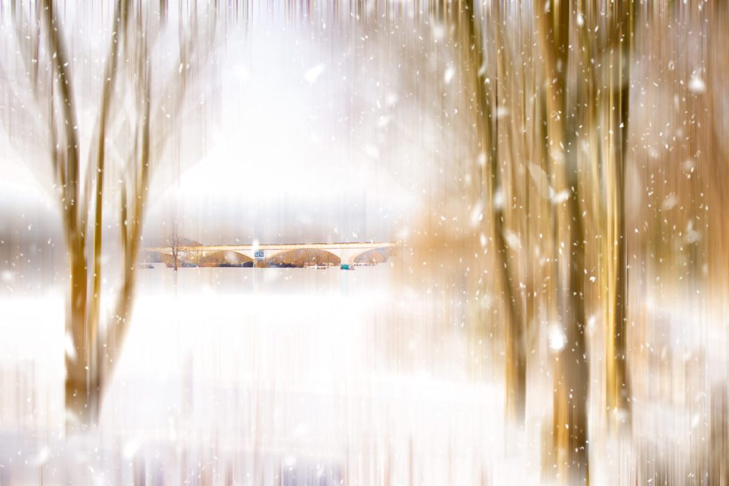Eine abstrakte, vertikal verwischte Fotografie einer Winterlandschaft. Im Zentrum ist eine ferne Brücke erkennbar, die von unscharfen, braunen Baumstämmen eingerahmt wird. Weiße Lichtpunkte, die an Schneeflocken erinnern, liegen über der gesamten hellen Szenerie.