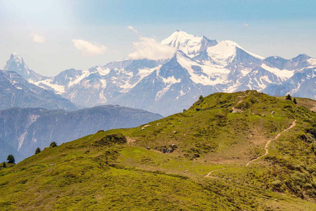 Ein grüner Berghang mit einem schmalen Pfad, Felsen und Grasbüscheln; dahinter ragen schneebedeckte, wolkenumhüllte Gipfel unter blauem Himmel auf.