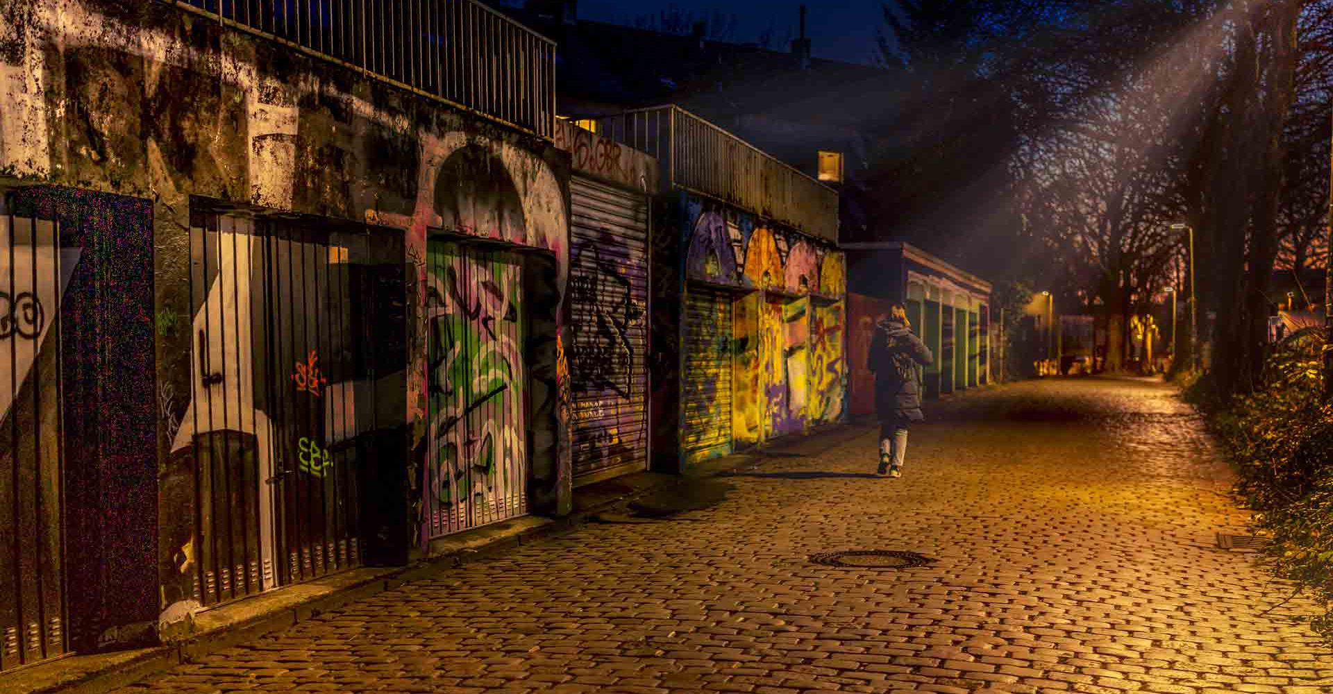 Person geht nachts eine Kopfsteinpflastergasse entlang, flankiert von bunt besprühten Garagentoren; warmes Straßenlicht und ruhige Atmosphäre.