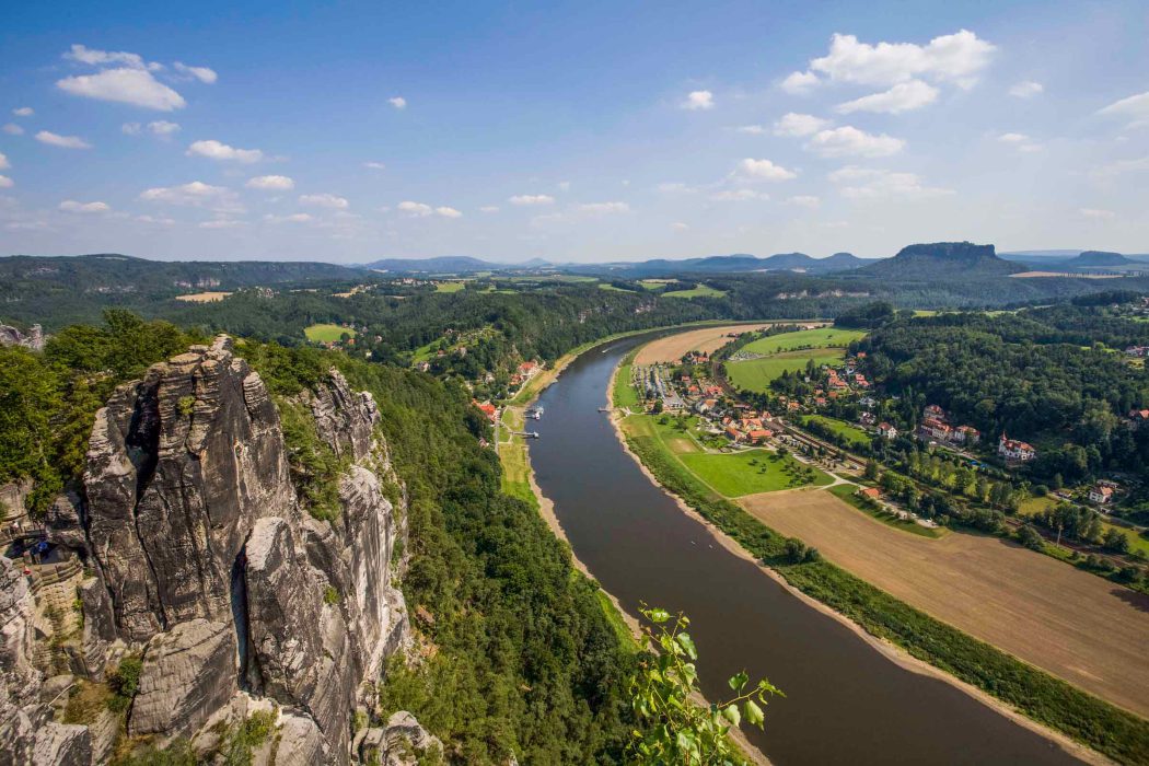 Panoramablick auf die Elbe, die sich durch ein Tal schlängelt, mit dramatischen Felsklippen links, grünen Wäldern, Feldern und einer kleinen Stadt unter einem teilweise bewölkten blauen Himmel.
