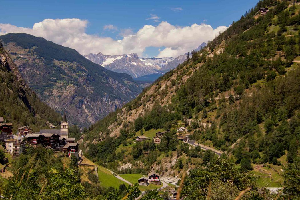 Panoramaaufnahme eines idyllischen Schweizer Bergdorfs in einem grünen Tal, umgeben von steilen Berghängen und schneebedeckten Alpen im Hintergrund. Traditionelle Chalets und eine Kirche mit Spitzdach prägen das Ortsbild, eingerahmt von satten Wäldern und klarer Gebirgsluft.
