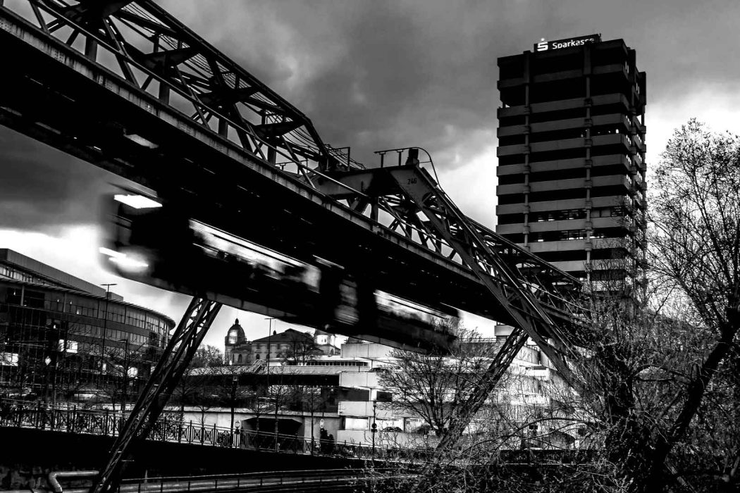 Schwarz-Weiß-Foto der Wuppertaler Schwebebahn in Bewegung mit dem Sparkassen Hochhaus im Hintergrund.