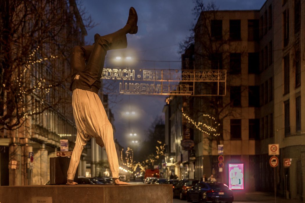 Kopfüber stehende Statue in einer Stadt bei Nacht mit beleuchteten Straßenschildern.