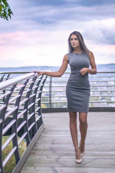ine schlanke Frau in einem eleganten, figurbetonten grauen Etuikleid und nudefarbenen High Heels steht auf einer modernen Holz- und Metall-Aussichtsplattform. Sie stützt sich leicht am Geländer ab und blickt nachdenklich in die Ferne über eine Stadtlandschaft unter einem bewölkten Himmel. Grüne Baumblätter sind im oberen linken Bildbereich sichtbar.
