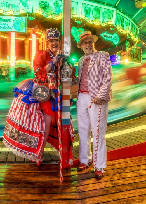 Zwei Personen in farbenfrohen, nostalgischen Kostümen stehen vor einem hell beleuchteten Fahrgeschäft; eine Figur im rot‑weiß‑blauen Outfit mit Zylinder, die andere im gestreiften Sakko mit Boater‑Hat und Zuckerstab.