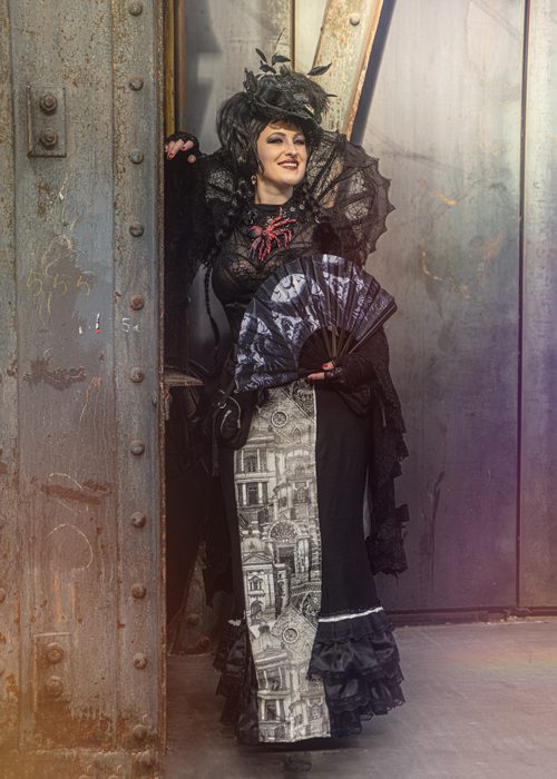 Person in detailreichem, schwarz‑viktorianischem Outfit mit Federkopfschmuck und Fächer steht vor einer industriellen Metalltür; Kontrast zwischen opulentem Kostüm und rauer Umgebung.