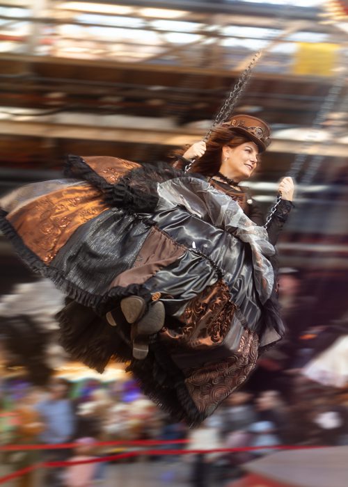 Person in aufwendigem historischen Kostüm schaukelt auf einer Indoor‑Jahrmarkt‑Schaukel; Bewegung, festliche Atmosphäre und detailreiche Kleidung stehen im Mittelpunkt.