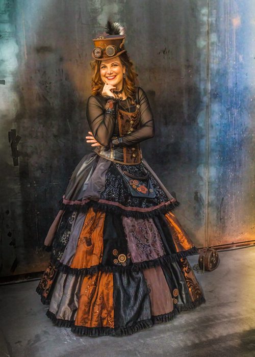 Person in aufwendigem Steampunk‑Kostüm mit langem, mehrlagigem Rock, Korsett, Hut und mechanischen Accessoires vor industriell anmutendem Hintergrund.