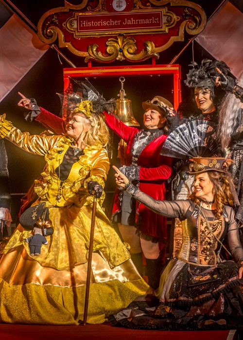 Gruppe von acht kostümierten Personen auf einer Bühne, in opulenten historischen und steampunk‑inspirierten Outfits, die gemeinsam in eine Richtung zeigen; festliche, theatralische Atmosphäre.