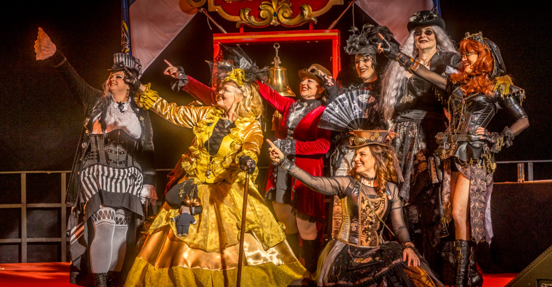 Gruppe von acht kostümierten Personen auf einer Bühne, in opulenten historischen und steampunk‑inspirierten Outfits, die gemeinsam in eine Richtung zeigen; festliche, theatralische Atmosphäre.