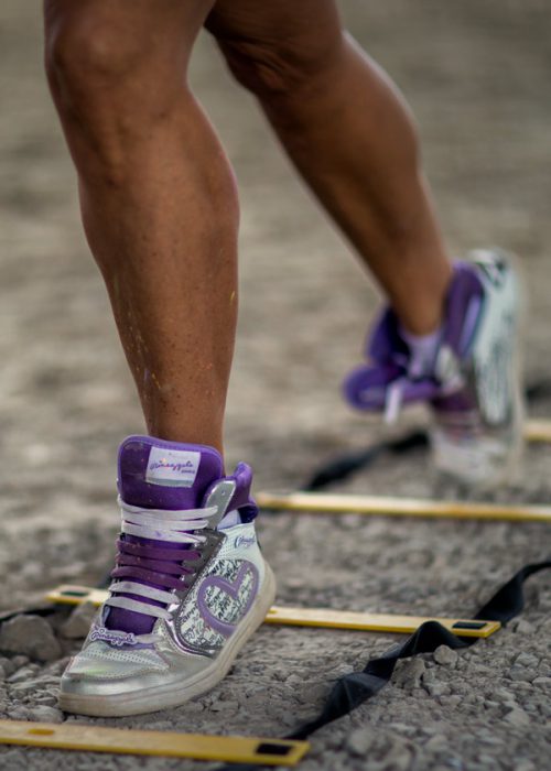 Die unteren Beine einer Person in lila-silbernen High-Top-Sportschuhen mit Schnürsenkeln sind zu sehen, wie sie über eine gelbe Koordinationsleiter auf einem Schotteruntergrund treten.