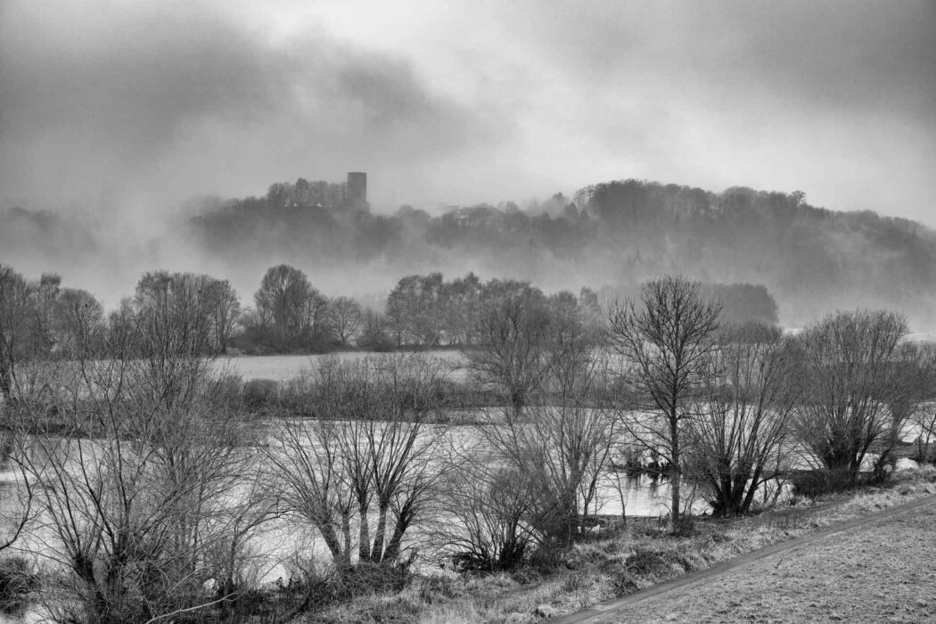 Schwarz-Weiß-Landschaft mit Fluss, kahlen Bäumen und nebliger Burg im Hintergrund