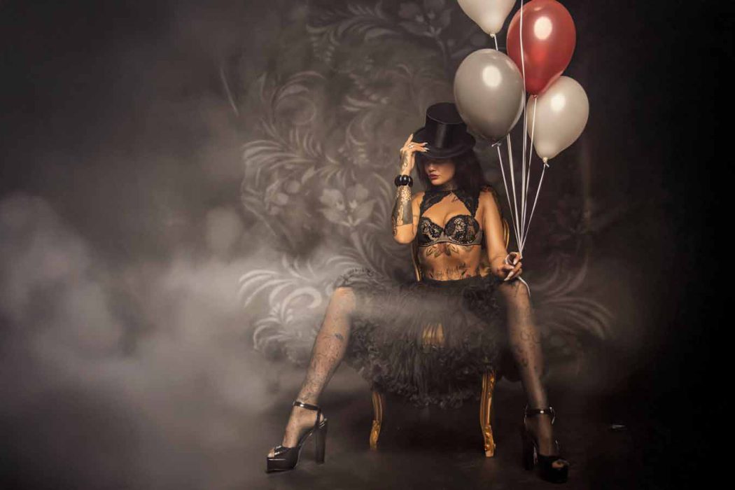 Kate mit einem Zylinderhut und Ballons posiert auf einem Stuhl vor einem dunklen, gemusterten Hintergrund mit Nebel. Die Person trägt ein Oberteil, einen Tüllrock und gemusterte Strumpfhosen. Boudoir Dotoshooting