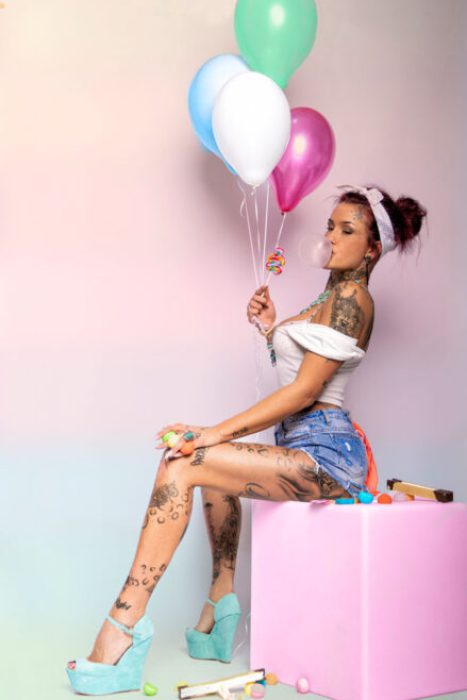 Eine junge Frau mit zahlreichen Tattoos an Armen, Beinen und Hals sitzt auf einem pinken Würfel. Sie trägt ein weißes Off-Shoulder-Top, Jeans-Shorts und mintgrüne Plateau-Wedges. In einer Hand hält sie drei bunte Ballons (grün, blau, pink) und eine Lutscher-Kette, während sie Kaugummi bläst. Um sie herum liegen bunte Süßigkeiten.