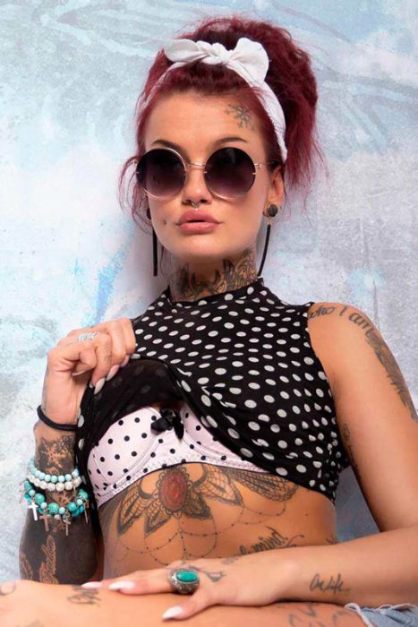 Ein Porträt einer Frau mit dunklem Haar, das in einem Dutt mit einem weißen Haarband hochgesteckt ist. Sie trägt eine runde Sonnenbrille und hat zahlreiche Tattoos auf ihren Armen, ihrem Hals und ihrem Bauch, sowie Piercings im Gesicht. Sie trägt ein schwarz-weiß gepunktetes Crop-Top über einem rosa gepunkteten BH und hat mehrere Armbänder und einen großen Ring an ihrer Hand.