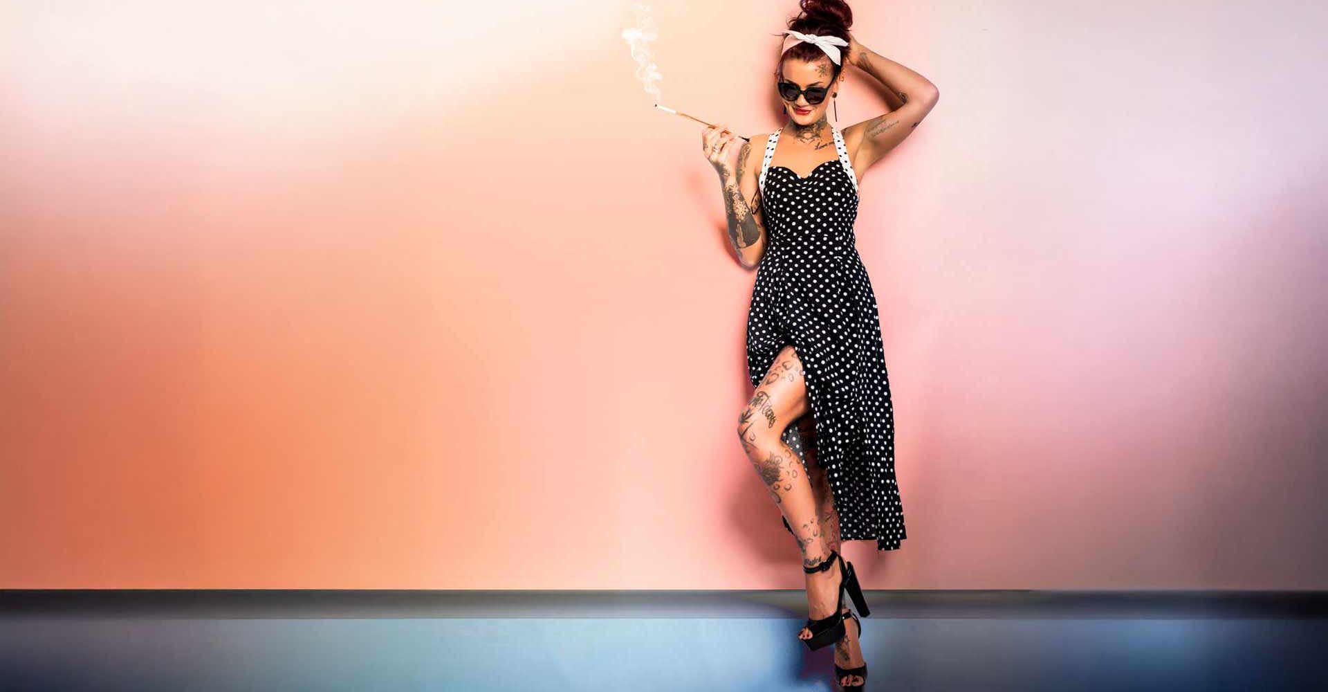 Ganzkörperaufnahme einer Frau im Vintage-Pin-up-Look: schwarzes Neckholder-Kleid mit weißen Punkten und Beinschlitz, Tattoos an Armen und Beinen, Sonnenbrille, Kopftuch und Zigarettenspitze, vor einer pastellrosa Wand mit dunklerem Bodenbereich