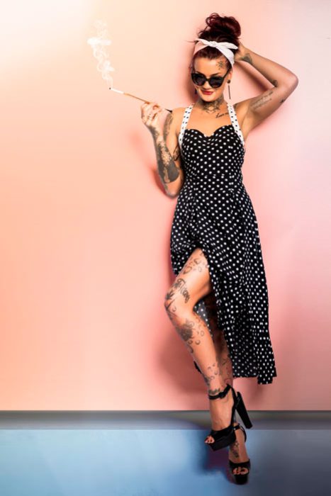 Ganzkörperaufnahme einer Frau im Vintage-Pin-up-Look: schwarzes Neckholder-Kleid mit weißen Punkten und Beinschlitz, Tattoos an Armen und Beinen, Sonnenbrille, Kopftuch und Zigarettenspitze, vor einer pastellrosa Wand mit dunklerem Bodenbereich
