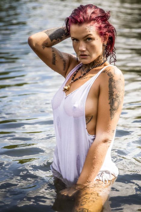 Porträt einer Frau mit roten Haaren und vielen Tattoos, die in einem See steht und ein nasses weißes Tanktop trägt.