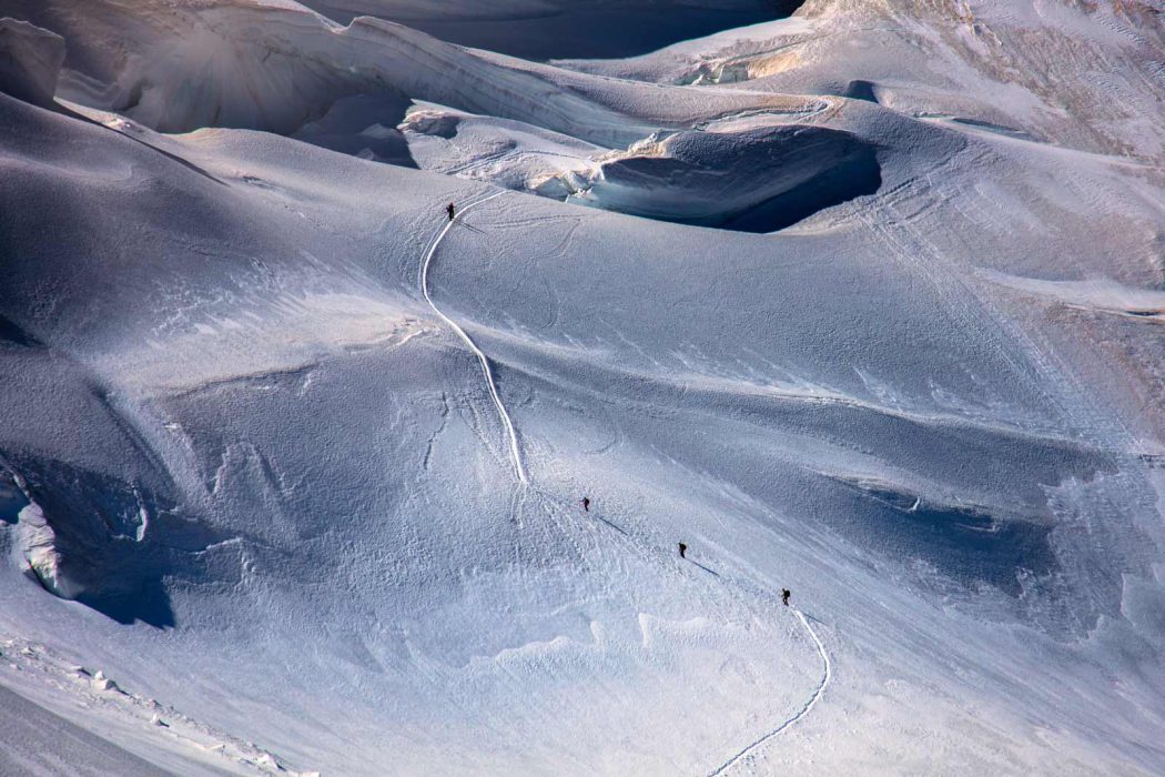 Luftaufnahme von Bergsteigern auf einem verschneiten Gletscherpfad in einer hochalpinen Gebirgslandschaft. Die kleine Gruppe folgt einer schmalen Spur durch das eisige Terrain mit sichtbaren Gletscherspalten und schroffen Schneestrukturen – ein eindrucksvolles Beispiel für alpines Abenteuer und Naturgewalt.