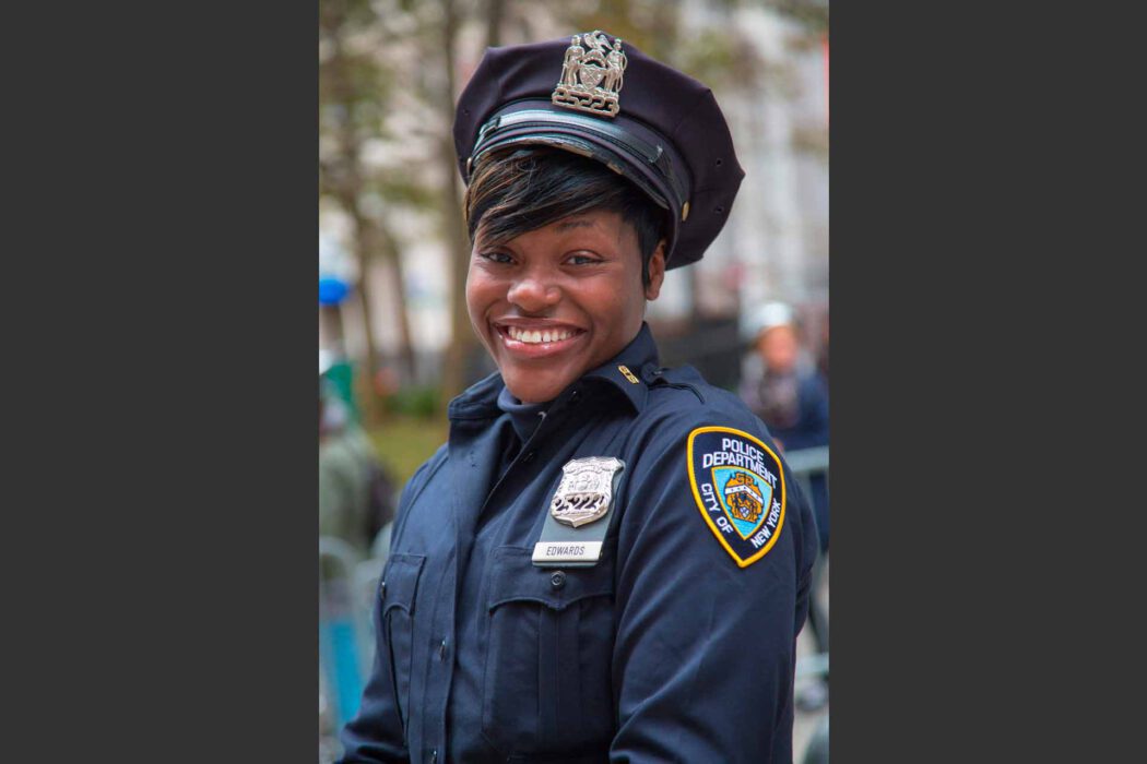 Reisefotografie New York: Lächelnde Beamtin des NYPD Police Department City of New York in dunkelblauer Uniform mit Mütze und Abzeichen, Porträt im Freien, unscharfer Hintergrund mit Bäumen, authentische Streetfotografie USA.