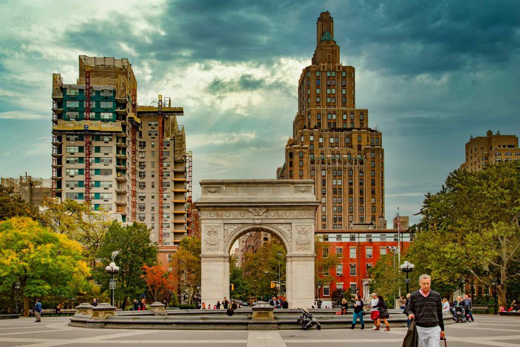 Dieses Bild zeigt den berühmten Washington Square Arch, das zentrale Wahrzeichen des Washington Square Park in New York City, unter einem teilweise bewölkten Himmel. Der prächtige Triumphbogen steht im Vordergrund, während im Hintergrund die beeindruckende Architektur der umliegenden Gebäude, darunter auch Gebäude der New York University, zu sehen ist. Der Park ist belebt mit Spaziergängern und verschiedenen Aktivitäten, was die lebendige Atmosphäre des Greenwich Village widerspiegelt. Bäume mit herbstlichem Laub und urbane Details vervollständigen die Szene. Dieses Bild ist ideal für Suchanfragen zu New York City, Parks, Sehenswürdigkeiten, Universitätsvierteln, urbanen Landschaften und Reisefotografie.