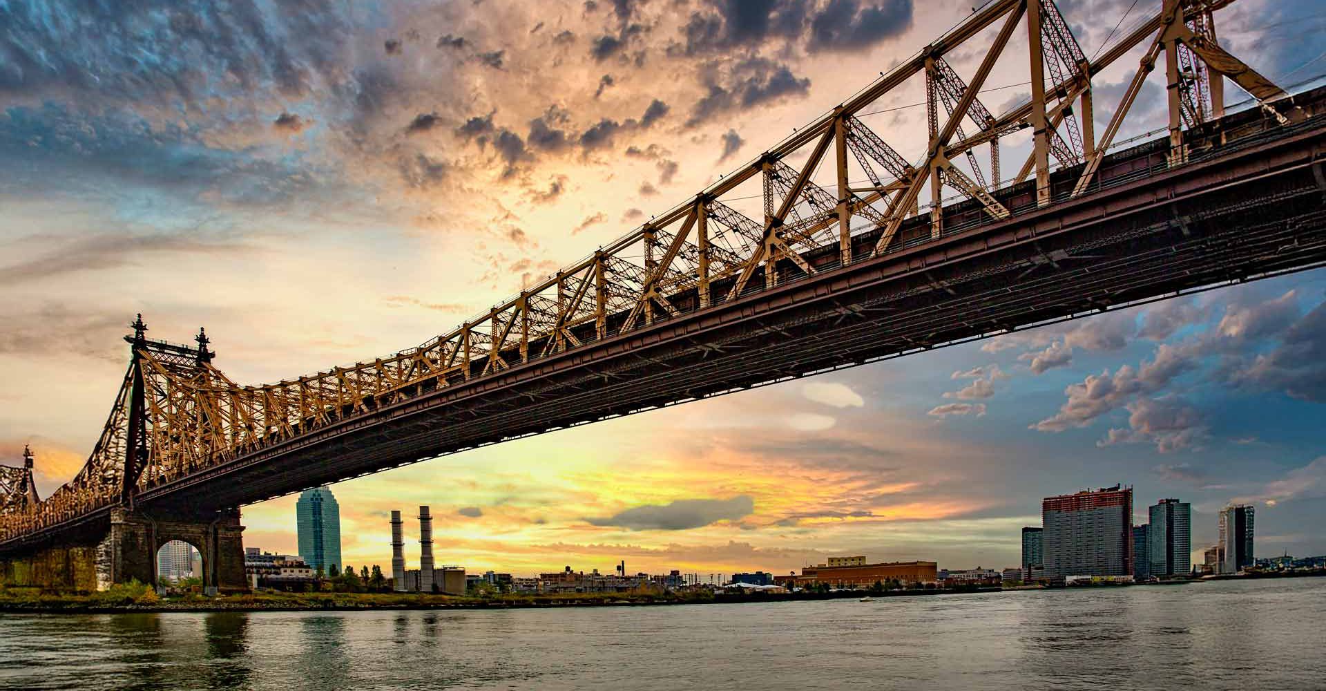 Queensboro Bridge bei Sonnenuntergang: Eine atemberaubende Ansicht der iconic Queensboro Bridge, die sich über den East River erstreckt, mit der Skyline von Manhattan und Long Island City im Hintergrund, alles getaucht in die warmen Farbtöne eines Sonnenuntergangs.