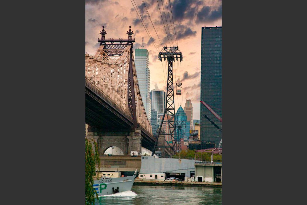 Dieses dynamische Bild zeigt zwei ikonische New Yorker Bauwerke: die majestätische Queensboro Bridge auf der linken Seite und die charakteristische Roosevelt Island Tramway in der Mitte. Eine der roten Tramway-Kabinen ist deutlich zu erkennen, wie sie über den East River schwebt. Im Hintergrund erheben sich moderne Wolkenkratzer der New Yorker Skyline, die einen starken Kontrast zu den historischen Strukturen bilden. Im Vordergrund ist ein Schiff, die "RED HOOK", auf dem Wasser zu sehen, was die maritime Umgebung unterstreicht. Der Himmel ist mit dramatischen Wolken versehen, die eine stimmungsvolle Atmosphäre schaffen. Dieses Bild ist ideal für Suchanfragen zu New York City, Brücken, Seilbahnen, urbaner Transport, Architektur und berühmten Sehenswürdigkeiten.