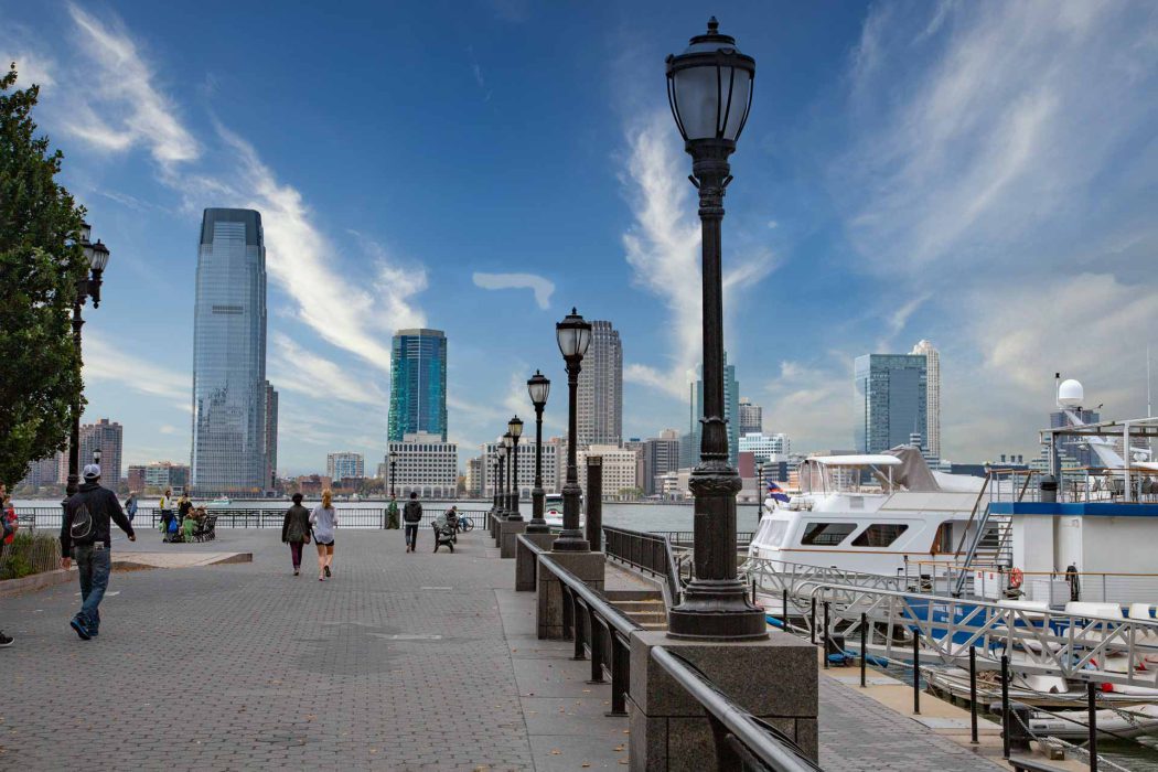 Dieses Bild zeigt die malerische Promenade des Battery Park in New York City an einem teilweise bewölkten Tag. Im Vordergrund erstreckt sich ein gepflasterter Weg mit klassischen Laternenmasten und einem Geländer, das zum Wasser hin abfällt. Entlang des Weges sind mehrere Personen zu sehen, die spazieren gehen oder die Aussicht genießen. Rechts sind Yachten und Boote an einem Pier vertäut. Im Hintergrund erhebt sich die imposante Skyline von Lower Manhattan oder Jersey City mit verschiedenen Hochhäusern und modernen Wolkenkratzern, die sich unter einem weiten blauen Himmel abzeichnen. Dieses Bild ist ideal für Suchanfragen zu New York City Sehenswürdigkeiten, Waterfront Parks, Stadtpromenaden, urbanen Landschaften und Reisefotografie.