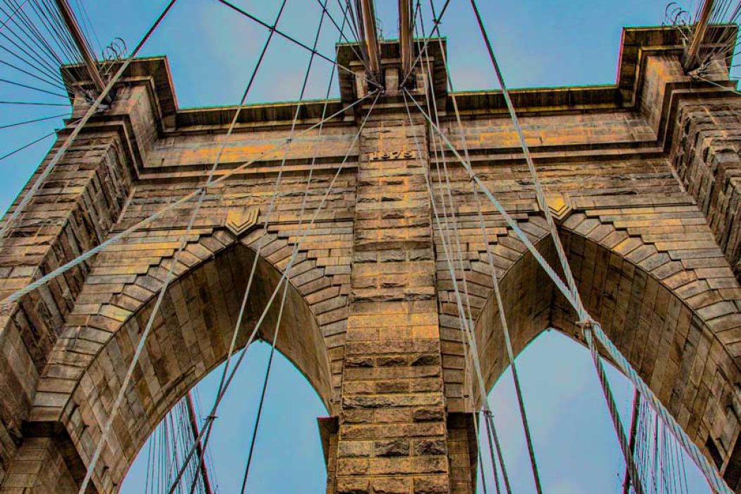 Dieses beeindruckende Nahaufnahme-Bild zeigt die massiven Steintürme und die markanten gotischen Bögen der berühmten Brooklyn Bridge aus einer Untersicht. Die detaillierte Steinmetzarbeit und die Kabel, die sich von den Türmen erstrecken, sind deutlich sichtbar und betonen die historische Ingenieurskunst und die majestätische Struktur des Bauwerks. Der helle, fast weiße Himmel im Hintergrund hebt die Silhouette der Brücke hervor und lässt ihre architektonische Pracht zur Geltung kommen. Dieses Bild ist ideal für Suchanfragen zu New York City, Architekturfotografie, berühmten Brücken, historischen Wahrzeichen und Ingenieurkunst.