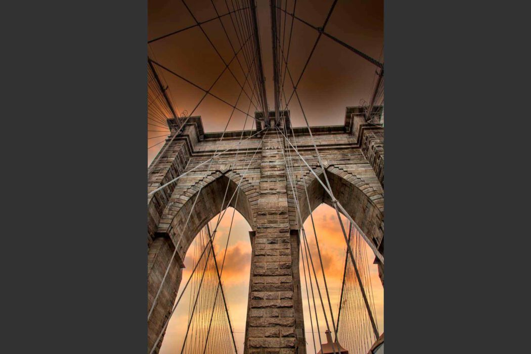 Steiler Blick auf den Steinpfeiler der Brooklyn Bridge mit Stahlseilen vor Sonnenuntergangshimmel.