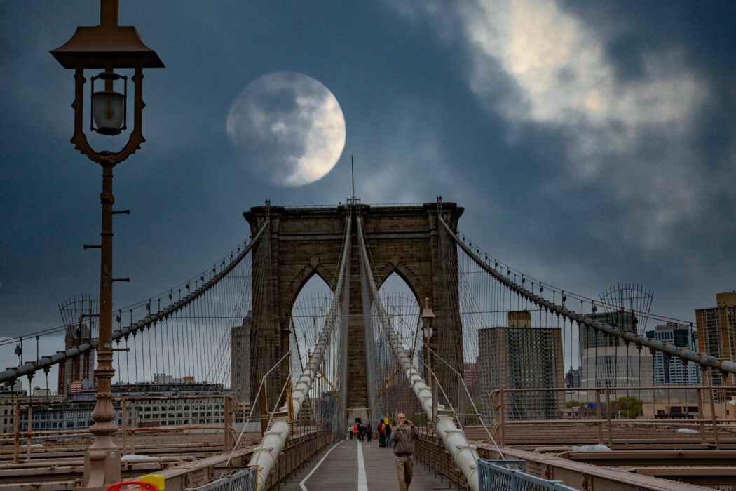 Reisefotografie Brooklyn Bridge New York bei Vollmond: Historischer Steinpfeiler mit gotischen Bögen, Stahlseile und Holzpromenade, große Laterne im Vordergrund, riesiger Mond hinter Wolken am dramatischen Himmel, Tourist fotografiert.