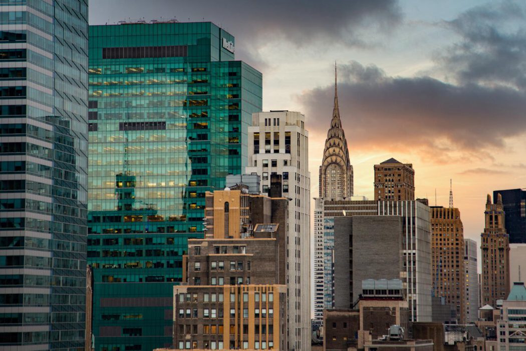 Reisefotografie Manhattan Skyline bei Sonnenuntergang: Chrysler Building mit Art-Deco-Spitze im goldenen Abendlicht, davor grünes MetLife Building mit Glasfassade, dichte Hochhäuser Midtown, dramatischer Wolkenhimmel.