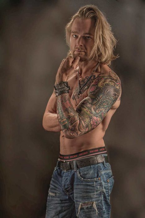 Ein Porträt eines muskulösen Mannes mit langen, blonden Haaren, der bis zur Taille abgebildet ist. Seine Arme sind vollständig mit farbigen Tattoos bedeckt, und er trägt mehrere Armbänder sowie eine Halskette. Er ist in zerrissenen Jeans gekleidet und seine rechte Hand ruht am Kinn, während er direkt in die Kamera blickt.