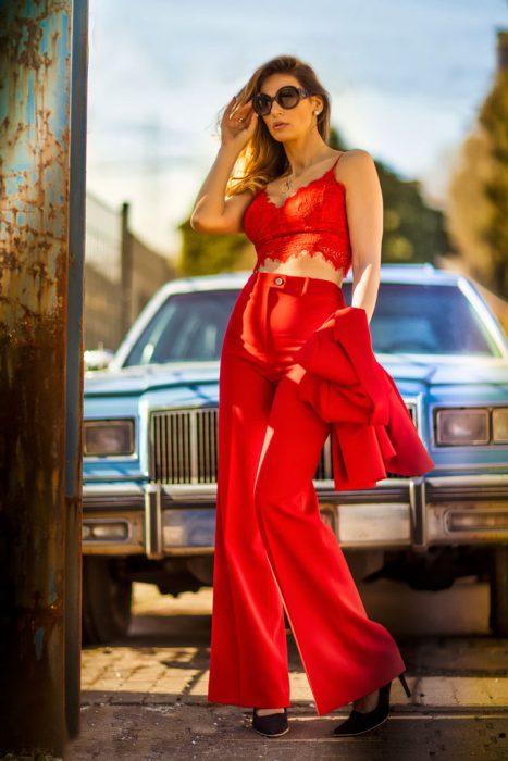 Eine stilvolle Frau trägt einen leuchtend roten Hosenanzug mit einem Spitzen-Crop-Top und hält eine passende Jacke über dem Arm. Sie steht selbstbewusst vor einem klassischen hellblauen Auto unter strahlendem Sonnenschein.