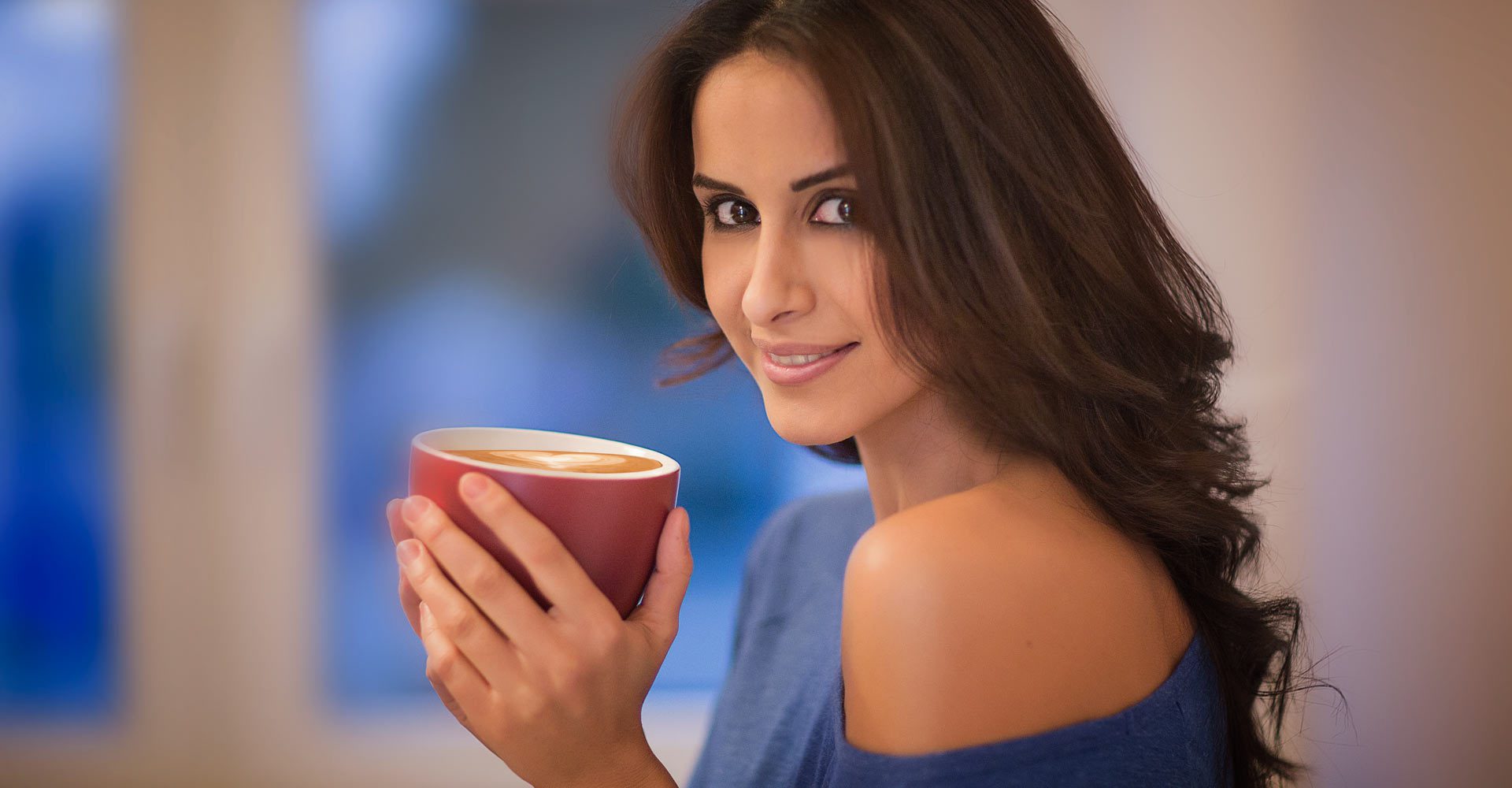 Person hält eine rote Tasse mit Latte‑Art in den Händen; weiches Licht, ruhige Innenraumatmosphäre, Fokus auf Geste und Wärme.