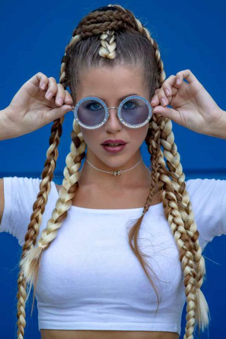 mit Zöpfen und Crop-Top, die eine runde Sonnenbrille vor einem blauen Hintergrund hält.