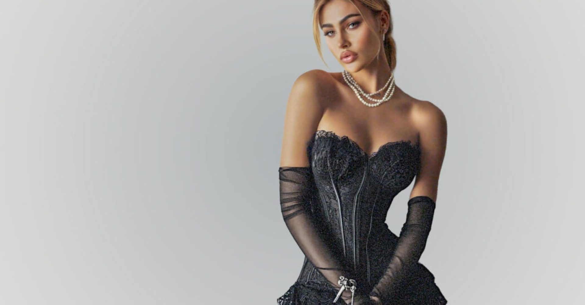 Eine Frau posiert in einem kunstvollen schwarzen Spitzenkorsett und langen, transparenten Handschuhen vor einem neutralen grauen Hintergrund. Sie trägt eine mehrreihige Perlenkette und hat ihr Haar elegant hochgesteckt.